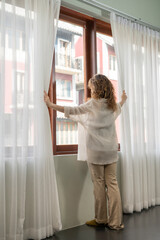 Fototapeta na wymiar The back shot of young woman open the white curtain for the morning sunshine. She stands by the window with the pose of open white sheer curtain with her arms. Lifestyle optimism concept