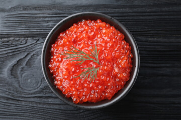 Bowl of delicious red caviar with dill on black wooden table, top view