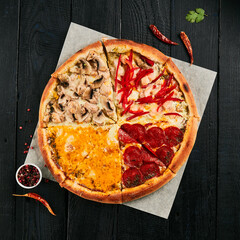Four seasons pizza on black wood background.  American pizza with pepperoni, mushrooms, tomatoes and cheese  on parchment. Italian food  in rustic style. Junk food on dark background