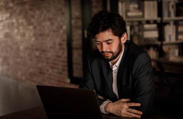 Caucasian successful professional diligent bearded and mustache male businessman in formal suit sitting at work desk typing laptop computer keyboard working late overtime in company office at night