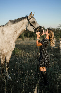 A young woman in a dress stands hugging and kissing her white horse in a field in the summer. Full-length photo, vertical, soft light, sunset. Love for animals, care for pets concept