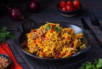 Pilaf, a dish of rice, chicken, carrots with spices and dried cranberries on a black plate on a dark wooden background.