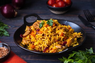 Pilaf, a dish of rice, chicken, carrots with spices and dried cranberries on a black plate on a dark wooden background.