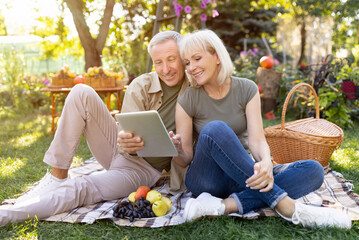Portrait of loving senior couple sitting on blanket during picnic and using digital tablet, resting outdoors