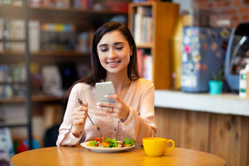 Young latin woman chatting on smartphone while eating fresh salad in cafe, using cellphone and surfing internet