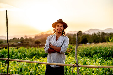 Young hipster farmer with long black hair standing arms crosses over a cornfield and sunset background. Attractive male with a sincere smile. Labor, happy hard work, hope, and rich concept ideas