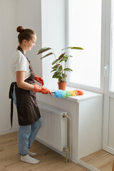 Side view full length portrait of woman housewife wearing brown apron and white t shirt cleaning her house, using colored ppduster for cleaning windowsill from dust.