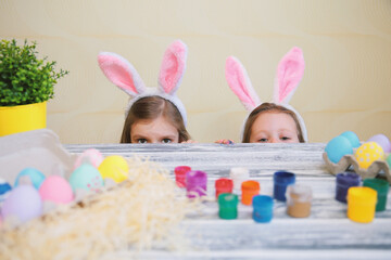 Curious child girls in bunny ears peeking out from behind table with colorful easter eggs. 