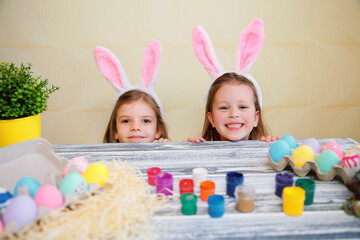 Funny child girls in bunny ears peeking out from behind table with colorful easter eggs. 