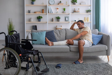 Unhappy black man having bandaged broken leg, sitting on sofa, suffering from pain at home, wheelchair standing nearby