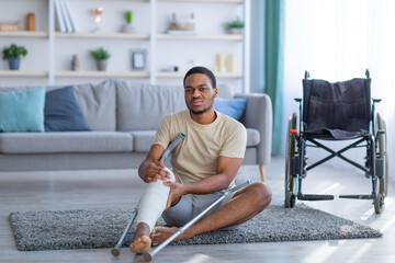 Fototapeta na wymiar Black guy sitting on floor after falling with crutches, suffering from pain in broken leg, wheelchair standing nearby
