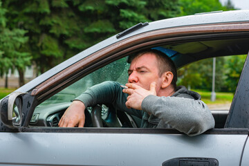 A man smokes in the rain while sitting in the car through the window