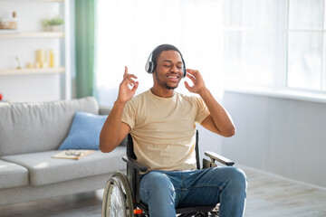 Cheery disabled black guy in wheelchair wearing headphones, listening to music with closed eyes at...