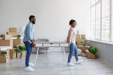 Cheerful young african american man and woman carry table in house with cardboard boxes, working together