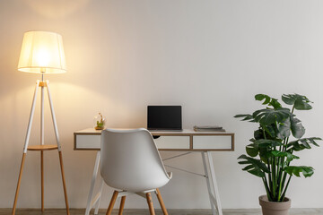 Domestic workplace with table, computer, armchair and glowing lamp, large potted plant on gray wall...