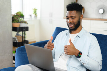 Cheerful multiracial guy taking a part in virtual meeting sitting with a laptop on laps on sofa at...