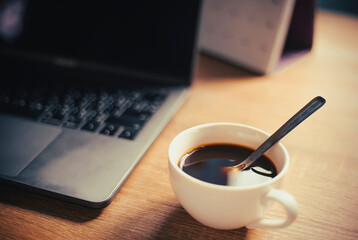 An image coffee hot drink on the white cup with a stainless spoon for breakfast with notebook or laptop is a beverage morning in office or home working cinematic style.