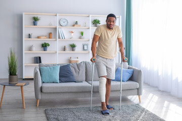 Young black guy with plastered leg walking with crutches, suffering from pain at home, copy space