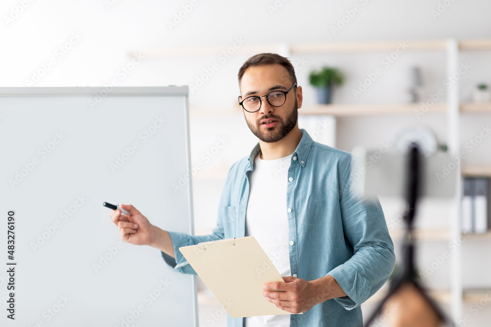 Poster young tutor giving online class, pointing at blank blackboard with mockup, explaining new material t