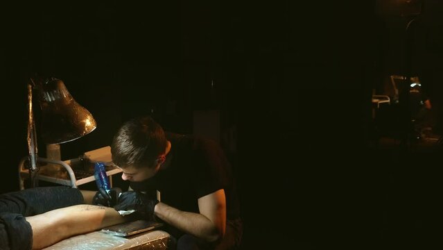 Young adult tattoo artist works with rotary machine on customers leg in dark lit room. Master tattooist with plastic film covered gun tattooing clients shin under lamp. Free space for advertisement.