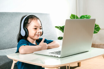 A  happy Little child girl studying online class on a laptop and headphones at home due to the Covid19 coronavirus pandemic. Online education learning gaming technology and homeschooling concept idea