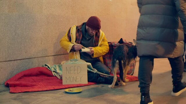 People are passing by a homeless man eating with his dog