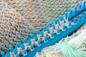 Obraz premium Colorful yarn texture background, beige creamy brown strings wrapped on pegs of long blue plastic loom. Shallow focus. In process of knitting warm soft winter clothes scarves and hats, home craft work