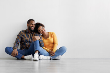 Satisfied young african american male and female sitting on floor on gray wall background, hugging,...