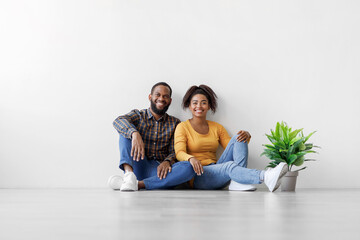 Glad young african american man and lady sit on floor with plant in pot on white wall background, rest after relocation