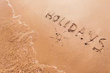 Holidays text with the sign of the sun on yellow sand on the beach by the sea. Summer vacation concept.