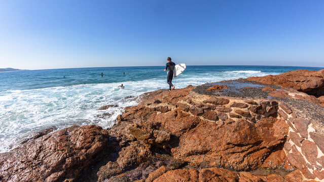 Surfer Going Surfing walking on rocky coastline point for jump entry into blue ocean waters a rear behind photo