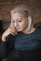 blonde girl in a sparkling sweater and a black corset sits on a chair against a brick wall in a loft