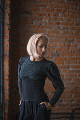 blonde girl in a shiny sweater, black corset and black trousers stands near a brick wall