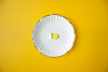 Top view of plate with two pills: yellow round tablet and white-yellow capsule. Yellow background and space for text. 