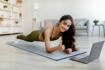 Obraz premium Online sports. Millennial Indian woman standing in elbow plank in front of laptop, showing thumb up gesture at home