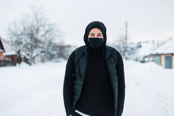Fototapeta na wymiar handsome young stylish hipster man with a protective mask in black fashionable outerwear with a hoodie, vest, hat and backpack walks on a winter day with snow. Guy in the town. Coronavirus concept