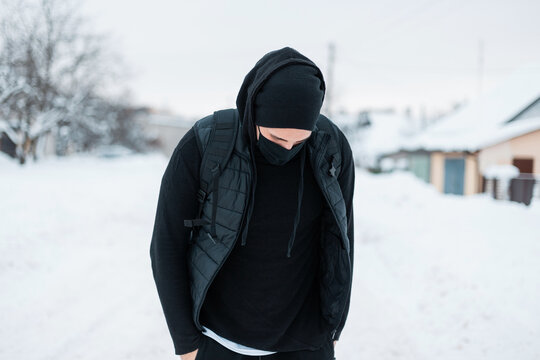 Fashionable handsome young hipster man in fashion black sports outerwear with hoodie, vest, hat and backpack walks on a winter day with snow in the countryside