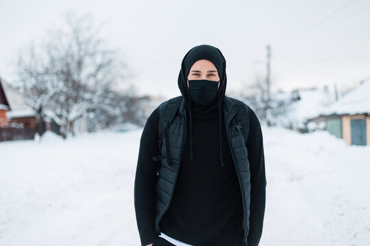 handsome young stylish hipster man with a protective mask in black fashionable outerwear with a hoodie, vest, hat and backpack walks on a winter day with snow. Guy in the town. Coronavirus concept