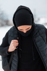 Stylish man with a black protective textile mask in fashionable black outfit with a backpack walks on the street against a background of snow on a winter day