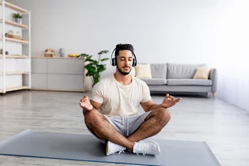 Fotobehang Peaceful young Arab man sitting in lotus pose, wearing headphones, meditating to relaxing music on mat at home © Prostock-studio