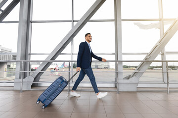 Portrait Of Smiling Arab Businessman With Suitcase Walking Near Window At Airport