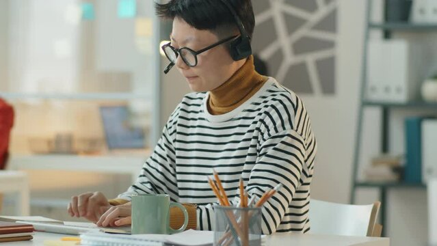 Tracking shot of Asian call center agent in wireless headset typing on computer and speaking on online call