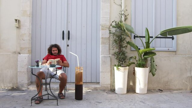 Timelapse of man in curly hair drinking a comically large iced coffee - frappe - outside a cafe.