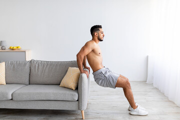Healthy living. Sporty young Arab guy doing strength exercises near sofa at home, copy space