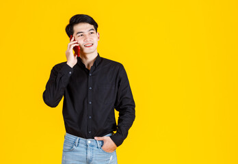 Studio shot of millennial Asian excited male businessman model in casual black long sleeve shirt and jeans standing calling smartphone on yellow background.