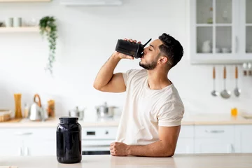 Poster Young Arab guy drinking protein shake from bottle at kitchen, copy space. Body care concept © Prostock-studio