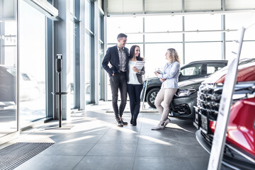 Young couple stands in teh car dealership discussing options with female shop assistant leaned against one of the cars