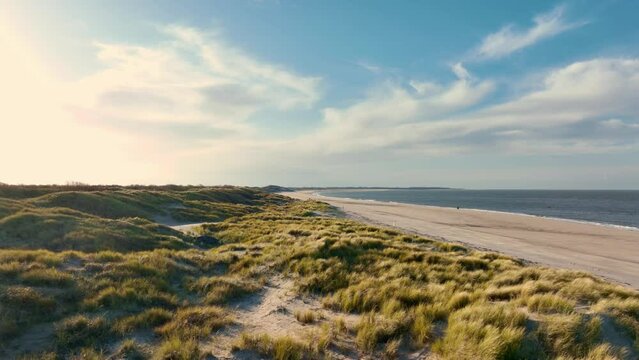 Long aerial shot flying over grassy dunes along an empty beach during a beautiful sunset