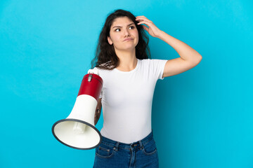 Teenager Russian girl isolated on blue background holding a megaphone and having doubts