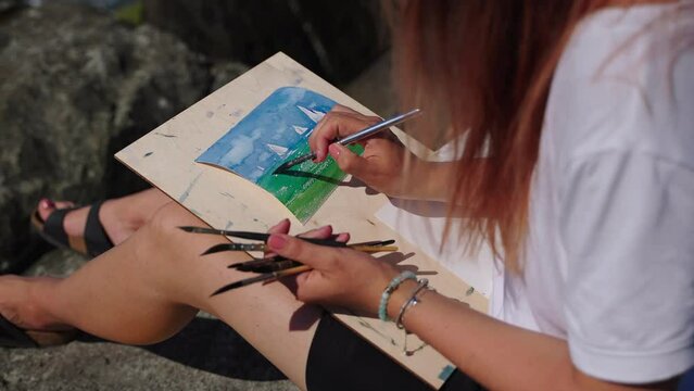artist woman is drawing seascape with sailboats, using aquarell dyes